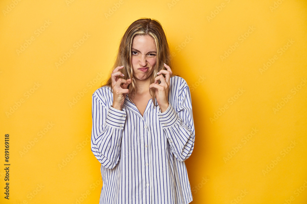 Young blonde Caucasian woman in a striped business shirt on a yellow background, upset screaming with tense hands. Young blonde Caucasian woman in a striped business shirt on a yellow background, upset screaming with tense hands.
