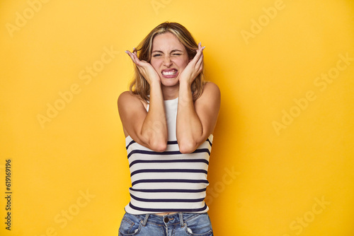 Young blonde Caucasian woman in a white tank top on a yellow studio background, covering ears with hands.