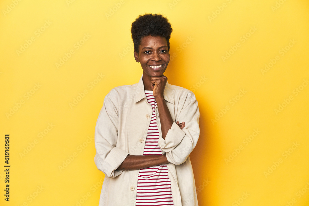 African woman, beige shirt, red striped tee, yellow studio, smiling happy and confident, touching chin with hand.
