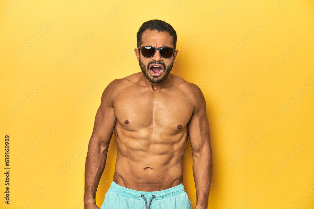 Fit young Latino man in swimwear and sunglasses, yellow studio background, screaming very angry and aggressive.