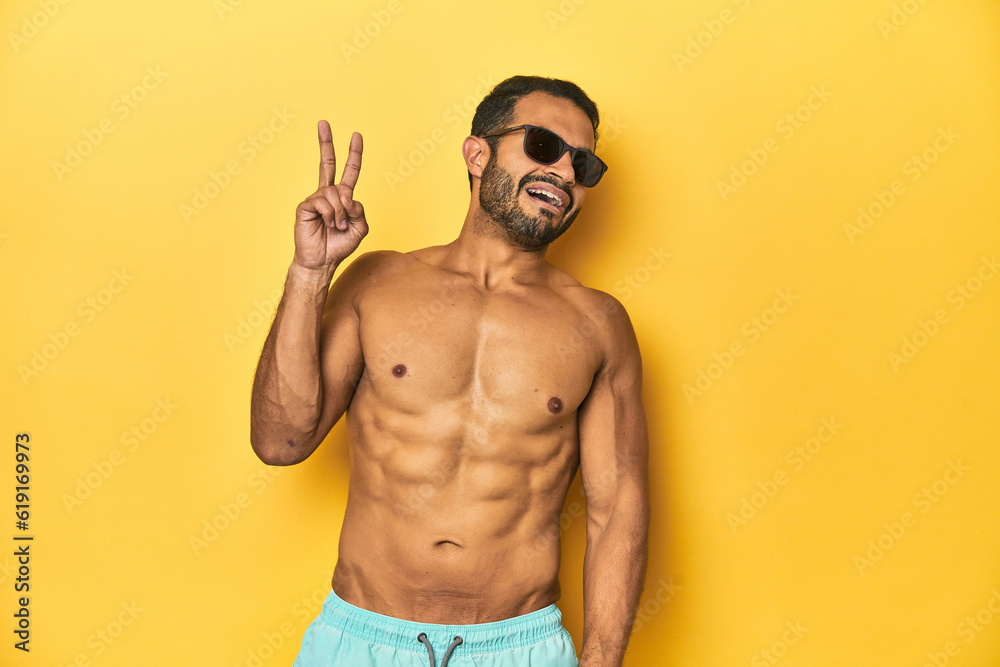 Fit young Latino man in swimwear and sunglasses, yellow studio background, joyful and carefree showing a peace symbol with fingers.
