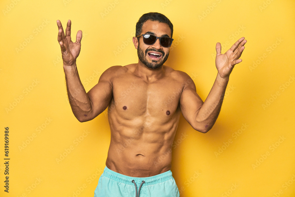 Fit young Latino man in swimwear and sunglasses, yellow studio background, receiving a pleasant surprise, excited and raising hands.