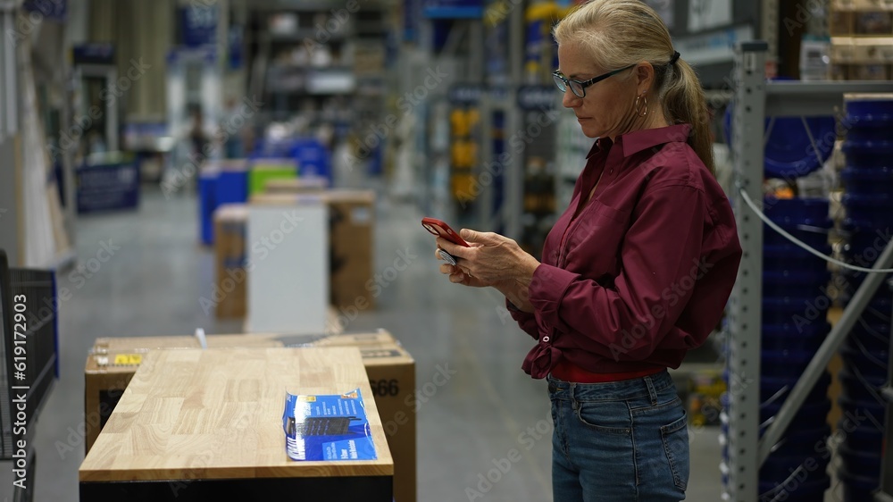 Mature woman uses phone to take photo of workbench furniture while shopping in large hardware store for home renovation project. Mature woman uses phone to take photo of workbench furniture while shopping in large hardware store for home renovation project.