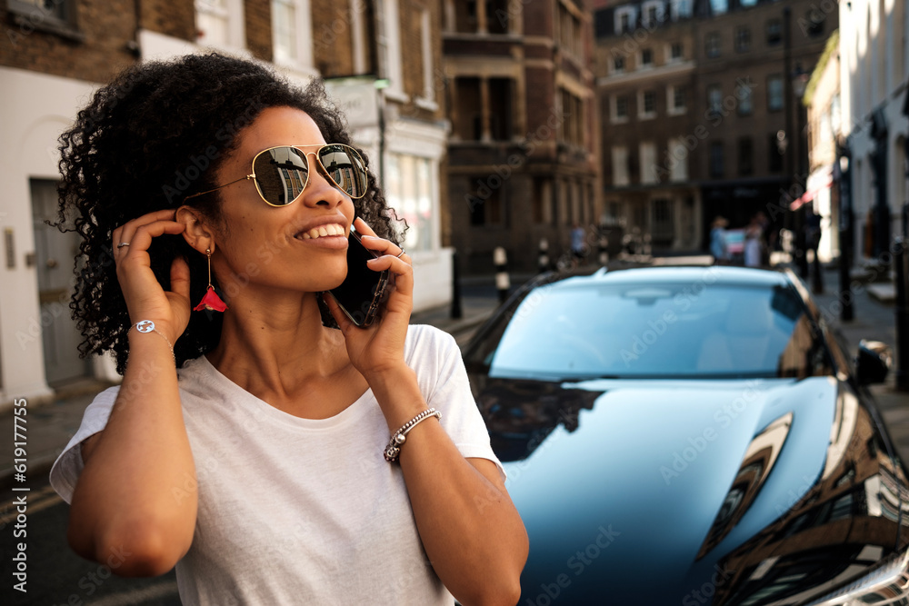Black smiling woman talking by phone in front luxury car.
