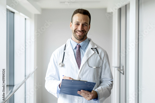  Portrait of a friendly european doctor in work clothes with a stethoscope around his neck, posing with folded hands in the interior of the clinic, looking and smiling at the camera, free space