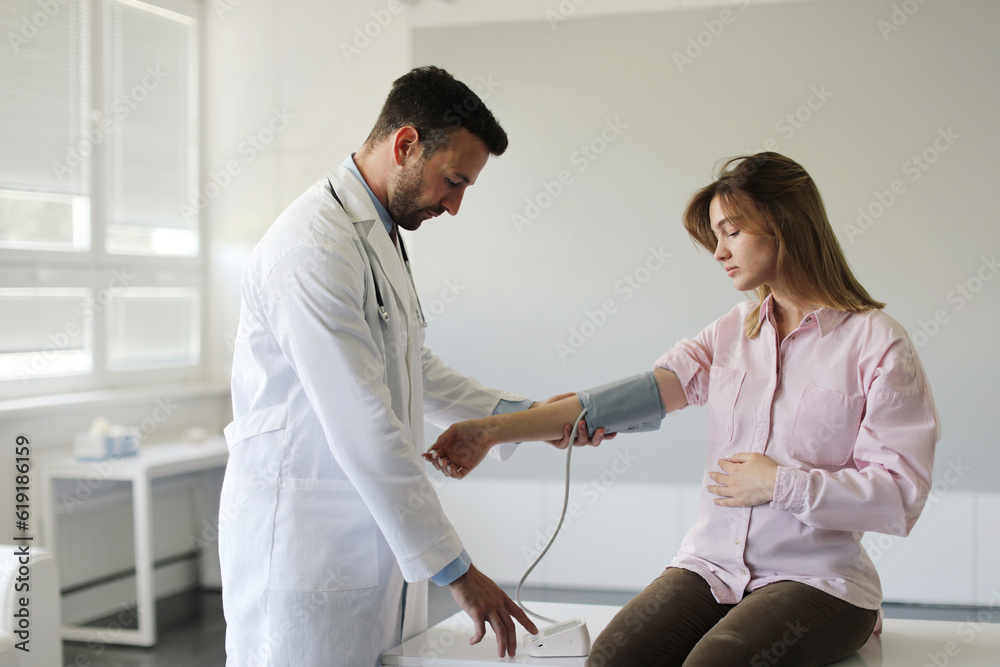 Obraz premium Confident man doctor checking young pregnant woman heart rate and blood pressure, using tonometer, working in clinic, copy space. Medical care and checkup during pregnancy