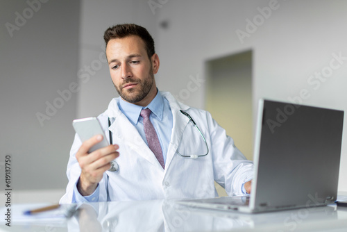 Focused male doctor in white uniform using cellphone and consulting patients online, sitting at desk with laptop in clinic office, copy space