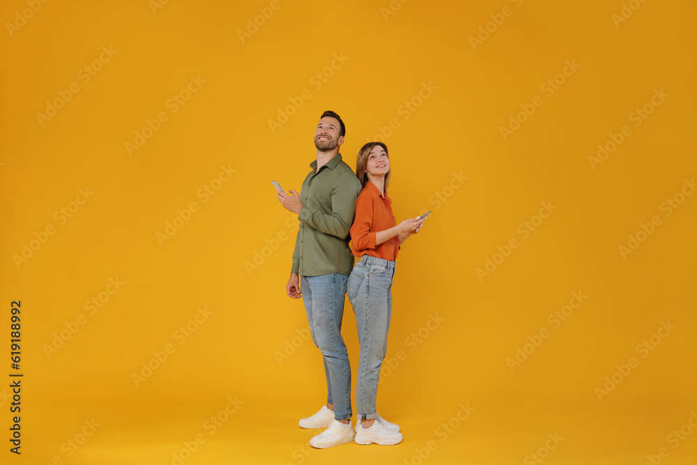 Full body length shot of two friends man and woman standing back to back, holding cellphones and looking up at free space, browsing internet isolated on yellow background Full body length shot of two friends man and woman standing back to back, holding cellphones and looking up at free space, browsing internet isolated on yellow background