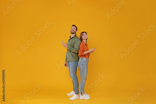 Full body length shot of two friends man and woman standing back to back, holding cellphones and looking up at free space, browsing internet isolated on yellow background