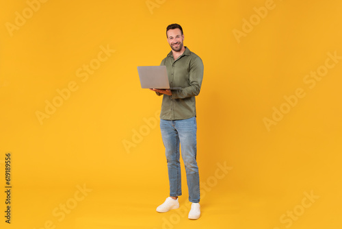 Happy european man with laptop for remote work or online shopping, posing over yellow studio background and smiling at camera, full length shot, copy space