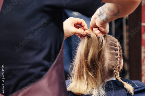 Close-up of hairstylist hands braiding braids for kid at barbershop. Hair salon, barber woman make hairstyle for pretty little blonde girl in barber shop. Hair care, beauty concept. Copy ad text space