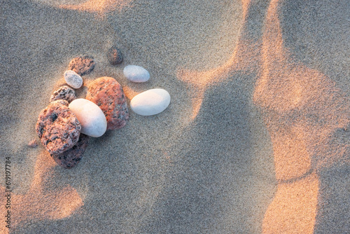 The uneven sand on the beach is illuminated by the soft light of the setting sun. There are several small sea pebbles of red and white color. Background. Texture.