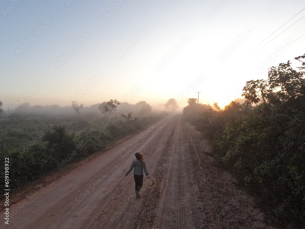 Amanhecer no Pantanal mato-grossense: Silhueta de pessoa não identificada com chapeu caminhadas na estrada transpantaneira em um amanhecer com neblina