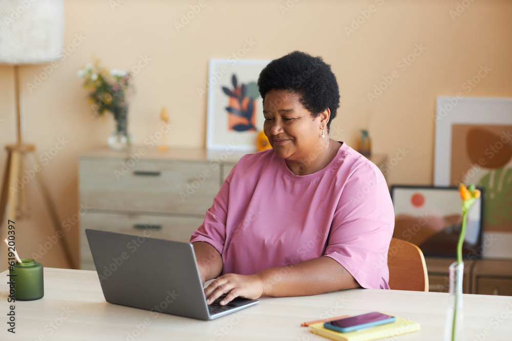 Happy mature African American woman in pink t-shirt sitting by desk in front of laptop in living room, typing on keyboard and networking Happy mature African American woman in pink t-shirt sitting by desk in front of laptop in living room, typing on keyboard and networking