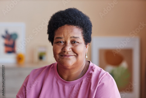 Mature African American grandparent in pink casualwear looking at camera while sitting in home environment against pictures on wall
