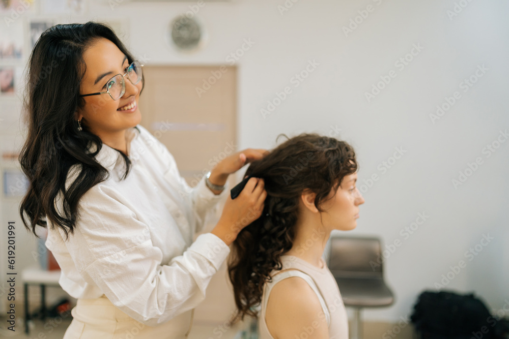 Side view of smiling female hairdresser combing long hair of young woman with hairbrush. Beautiful young woman getting haircut by hairstylist in beauty salon. Concept of fashion and beauty. Side view of smiling female hairdresser combing long hair of young woman with hairbrush. Beautiful young woman getting haircut by hairstylist in beauty salon. Concept of fashion and beauty.