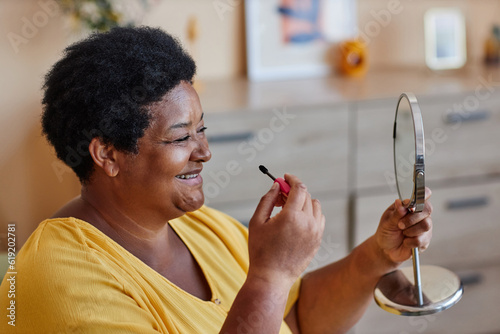 Mature African American laughing woman in yellow casualwear looking at herself in mirror and applying mascara on eyelashes in the morning