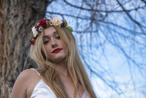 Portrait of a young, beautiful, blonde woman in a white dress and flower headband, posing next to a dry tree, calm and relaxed. Concept beauty, fashion, brides, peace, relaxation.