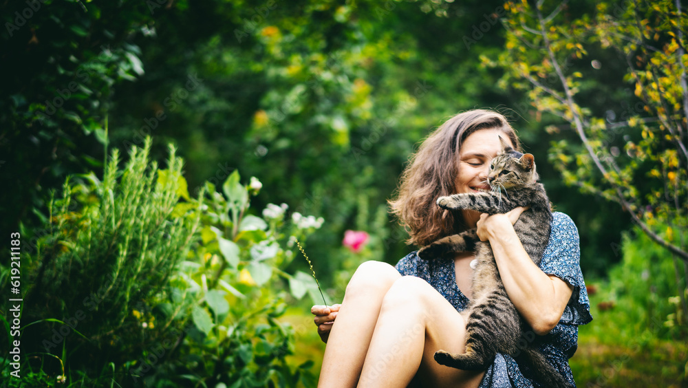 Young cheerful smiling woman holding in her arms a cute gray cat sitting on the grass in a summer garden
