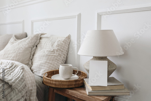 Closeup of blank greeting card, invitation mockup. Breakfast in bed concept. Cup of coffee, table lamp with linen shade, vintage wooden night stand. Checkered beige pillows, blurred background. © tabitazn