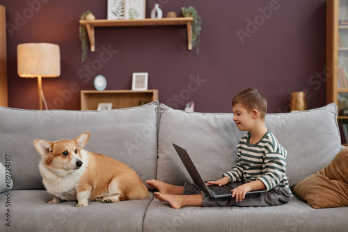 Side view portrait of curious little boy with down syndrome using laptop computer while sitting on comfortable sofa with dog, copy space