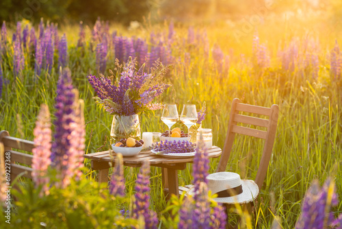 Romantic table decor for a loving couple on the blooming meadow with purple lupines. Two glasses of wine, flowers in a vase, silverware, fruits, wooden furniture and picnic basket. Sunset, golden hour