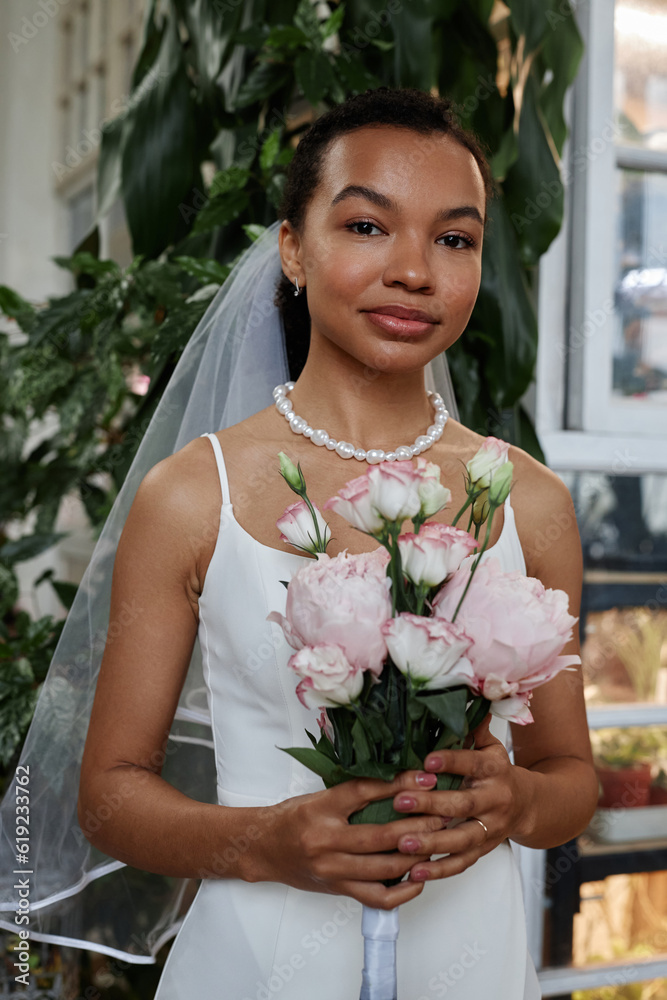 Vertical portrait of beautiful black woman as young bride wearing simple wedding gown and holding bouquet looking at camera