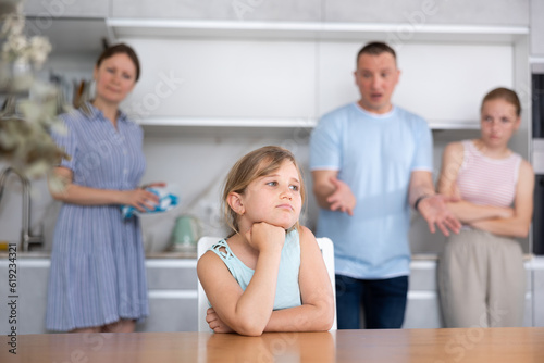 Portrait of frustrated tween girl in home kitchen, frowning with arms crossed. Disgruntled parents reprimanding her while standing with eldest daughter on blurred background. Emotional abuse concept