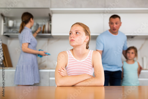 Upset teenage girl sitting at table in home kitchen, frowning and crossing arms. Discontented father reprimanding her while standing with younger daughter in blurred background, mother washing dishes