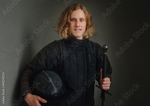 Portrait of a smiling long-haired young teen dressed in black historical fencing armor with a long medieval sharp sword