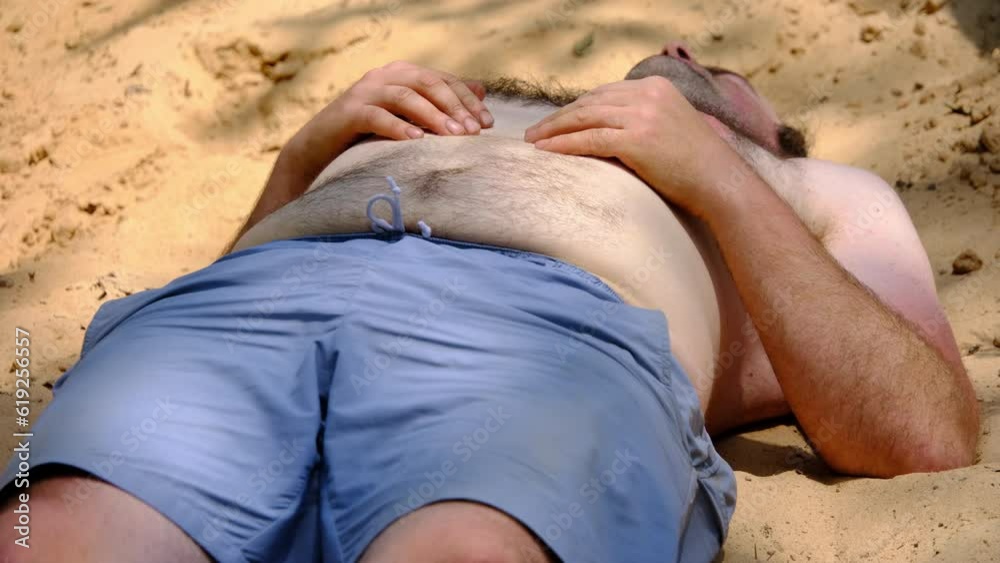 Close up of slightly obese man with blue shorts and topless with hairy chest waking up and stretching after falling asleep on top of pile of sand after back yard pool party.