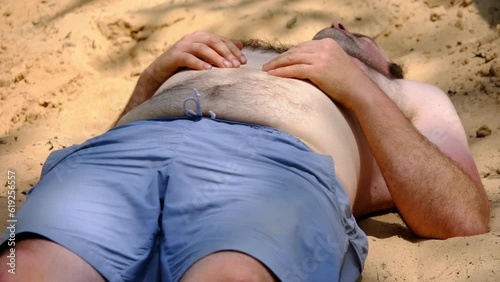 Close up of slightly obese man with blue shorts and topless with hairy chest waking up and stretching after falling asleep on top of pile of sand after back yard pool party. photo