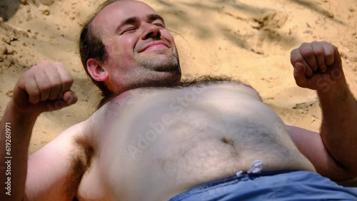 Close up of young man with blue shorts and topless with hairy chest waking up and stretching after falling asleep on top of pile of sand after back yard pool party. photo