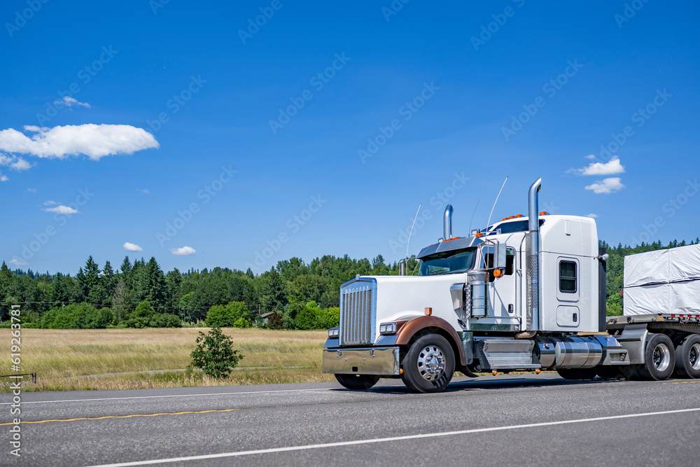 Powerful big rig American semi truck tractor transporting covered fastened cargo on flat bed semi trailer driving on the highway summer road at sunny day