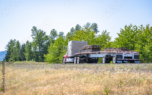 Powerful bonnet big rig beige semi truck transporting fastened by slings lumber wood on the flat bed semi trailer driving on the road in scenic nature area