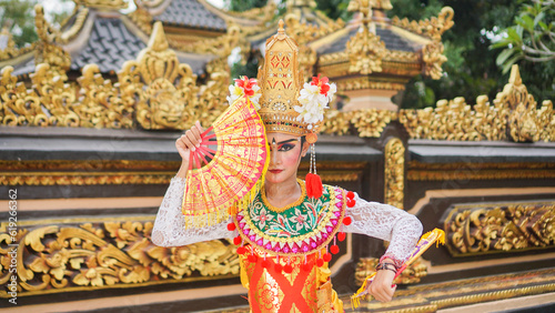 girl wearing Balinese traditional dress with a dancing gesture on Balinese temple background with hand-held fan, crown, jewelry, and gold ornament accessories. Balinese dancer woman portrait 