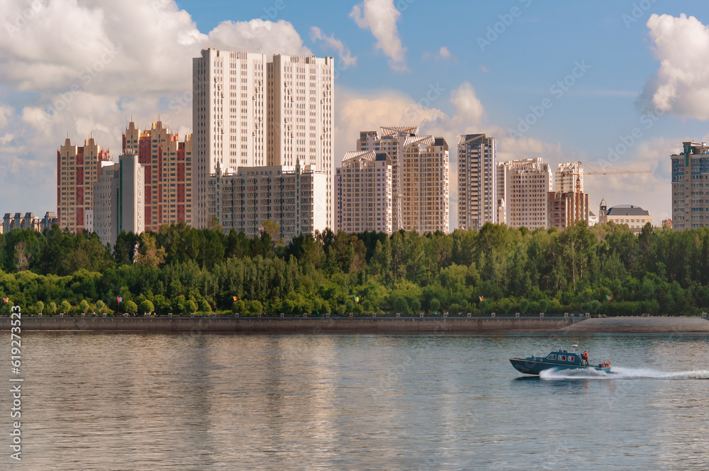 Buildings of a residential urban area on a wooded shore. A speedboat rushes along the surface of the water of the river. Blue sky with white clouds. Sunny summer evening at golden hour.