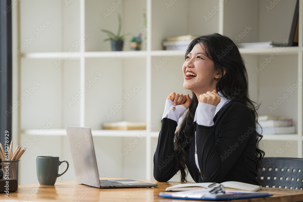 An Asian businesswoman celebrating victory success, an employee inside the office reading good news, using a laptop at work inside the office holding hand up, and a happy triumph gesture.