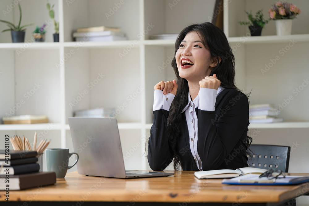 An Asian businesswoman celebrating victory success, an employee inside the office reading good news, using a laptop at work inside the office holding hand up, and a happy triumph gesture.