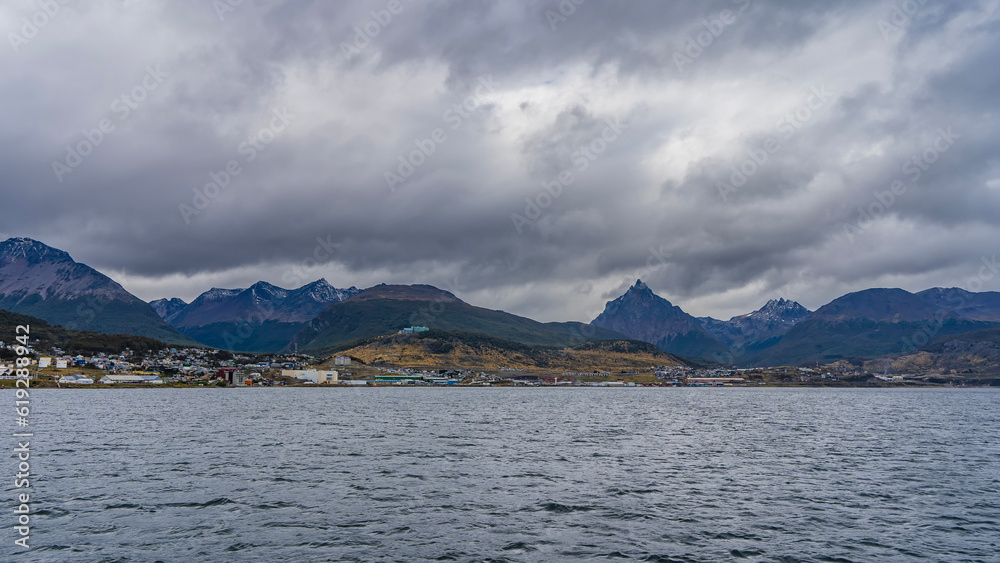 From the side of the Beagle Canal, a coastal strip with city houses is visible. A picturesque mountain range of the Andes against a cloudy sky. Ripples on the water. Ushuaia. Argentina.