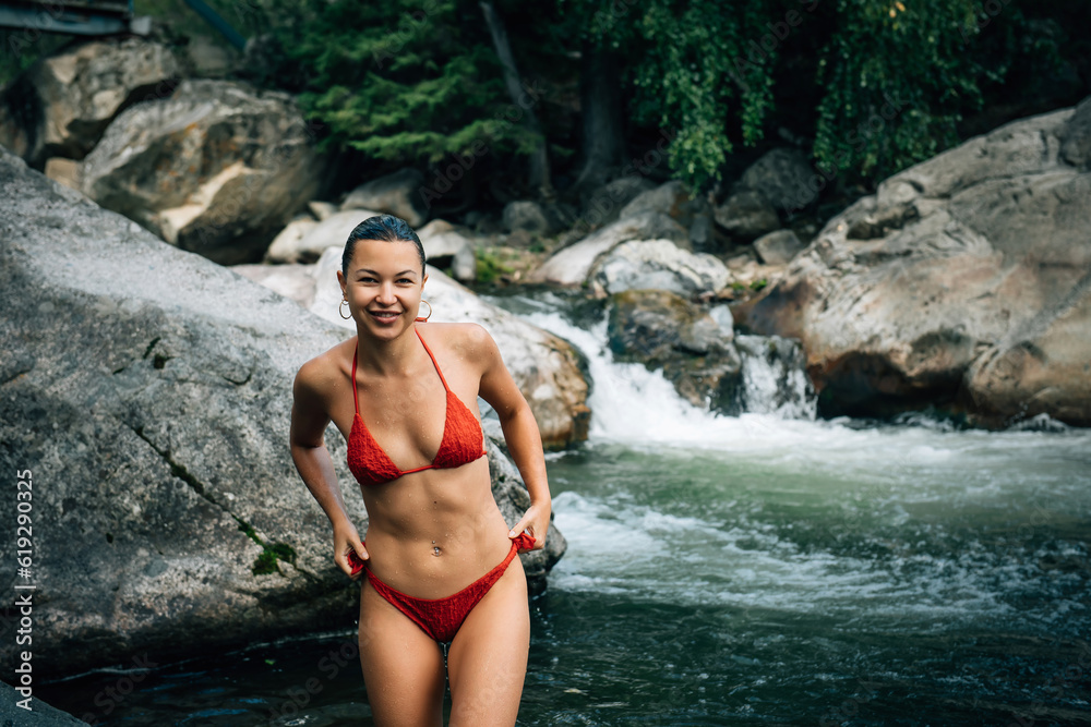 Beautiful young woman wearing red bikini coming out stormy mountain river. Happy female brunette with sexy body on picturesque natural background.