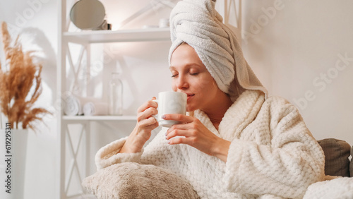 Morning coffee. Weekend relaxation. Spa leisure. Happy inspired woman in bathrobe with mug enjoying hot drink aroma on couch after bath at light home interior with free space. photo