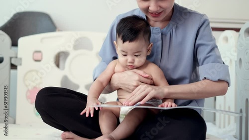 Happy Time, Asian Mother Reading a Storybook to Her Adorable 5-Month-Old Baby in a Playful Home Setting, Fostering Connection and Early Development.