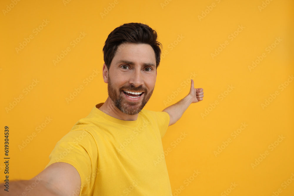 Smiling man taking selfie and showing thumbs up on yellow background, space for text Smiling man taking selfie and showing thumbs up on yellow background, space for text
