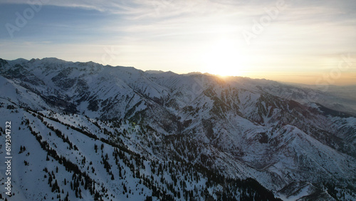 Epic red-orange sunset in high snowy mountains. There are tall coniferous trees, there is snow on the hills. High peaks. The sun's rays fall on the clouds and fog that is lower down the gorge. Almaty