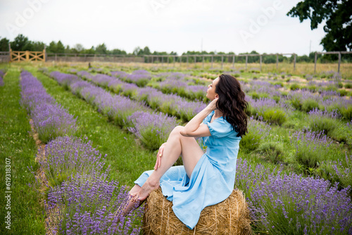 Fototapeta Naklejka Na Ścianę i Meble -  beautiful brunette smiling woman in blue dress sitting on the straw bale in lavender field on summer day
