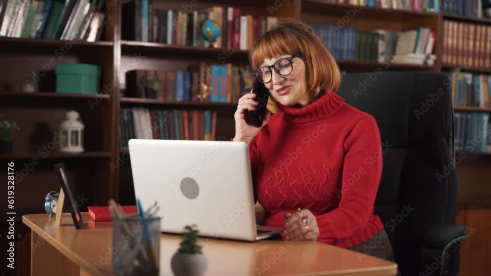 The woman is sitting at the table, holding a phone in her hands. She is talking to someone on the phone, and she appears to be happy. It is evident that she was working before she received the call. The woman is sitting at the table, holding a phone in her hands. She is talking to someone on the phone, and she appears to be happy. It is evident that she was working before she received the call.