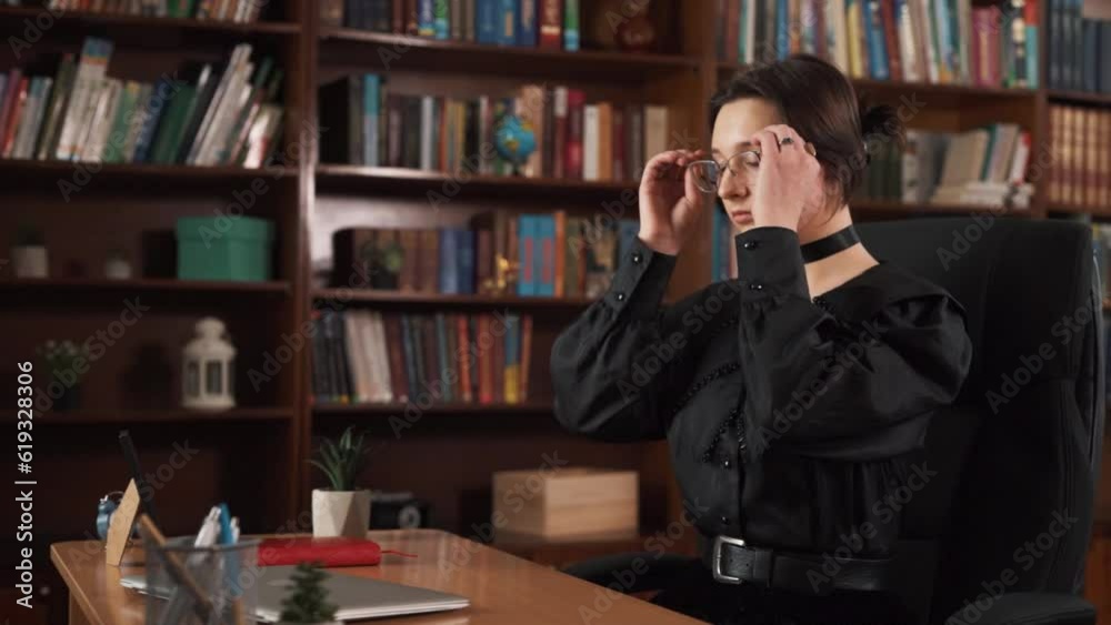 The girl is seated at a table, with a chair, and she has a laptop placed on it. Engaged in work, she is typing on the laptop. She puts on glasses and is surrounded by numerous books. The girl is seated at a table, with a chair, and she has a laptop placed on it. Engaged in work, she is typing on the laptop. She puts on glasses and is surrounded by numerous books.