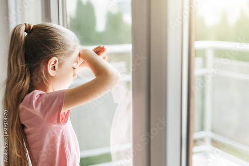 Sad little girl looking through window at home.