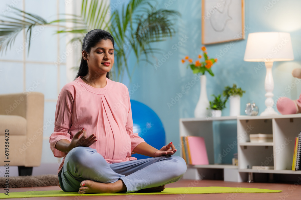 Indian peaceful pregnant woman in lotus pose doing meditation on yoga mat home - concept of mindfulness, relaxation and Self-care during pregnancy - obrazy, fototapety, plakaty 